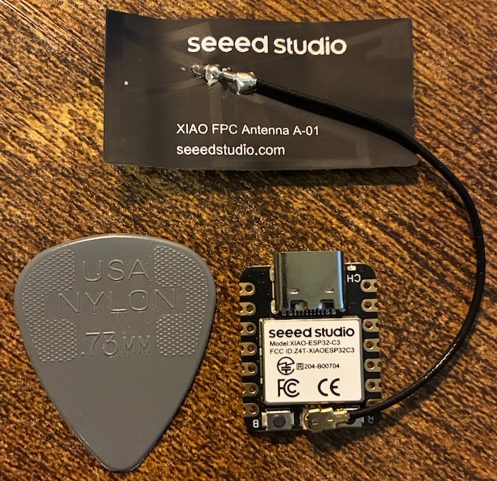 A Seeed Studio XIAO ESP32C3 next to a gray Jim Dunlop nylon 0.73 mm guitar pick, with the XIAO FPC antenna still on its backing card